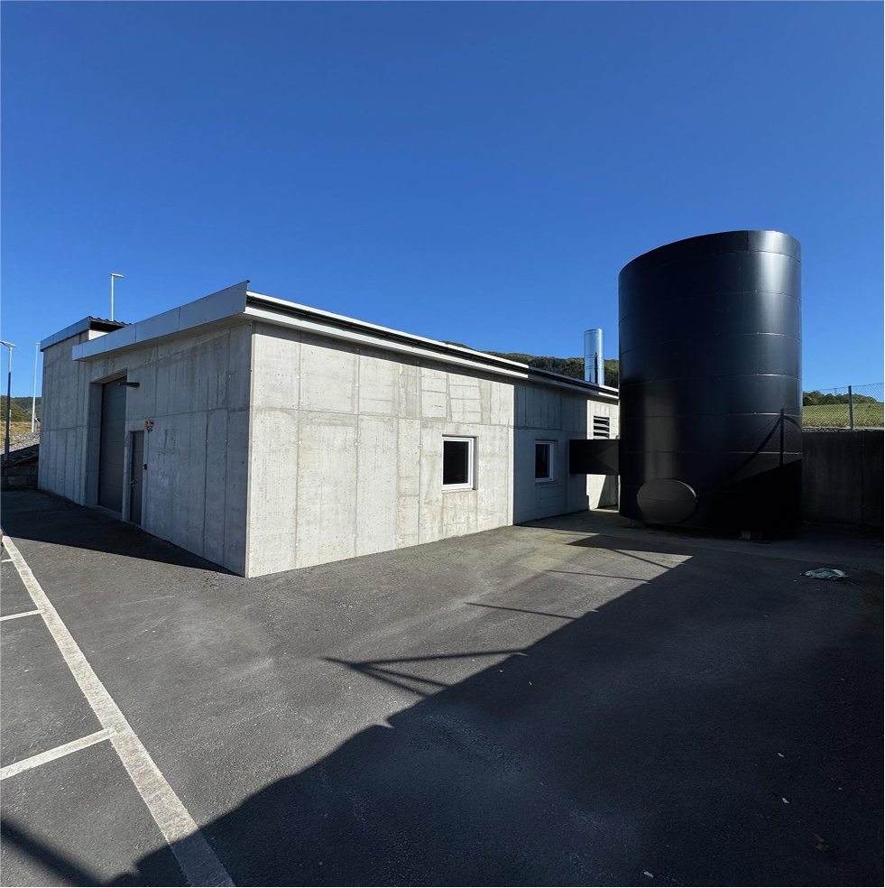 Moderne energisentral i betong med sort akkumulatortank for varme- og kjøleløsning, plassert ved fjellgrunn.